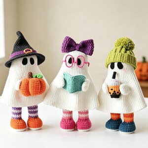 May include: Three crocheted ghost dolls. The first wears a black witch hat and holds an orange pumpkin. The second wears a purple bow and pink glasses while holding a teal book. The third wears a lime green pom-pom beanie and holds a boba tea.