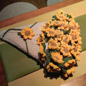 K&ouml;nnte beinhalten: Ein geh&auml;kelter Sonnenblumenstrau&szlig; in einem cremefarbenen Korb. Die Sonnenblumen sind gelb mit braunen Zentren und gr&uuml;nen Bl&auml;ttern.
