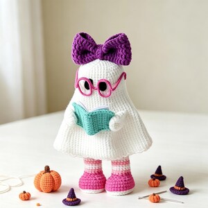 May include: A white crocheted ghost figure wearing a large purple bow, pink round glasses, and pink and white striped socks. The ghost holds a small teal book. Surrounding the figure are several small crocheted orange pumpkins and tiny purple witch hats.