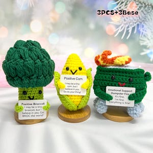 May include: Three crocheted vegetable figures: a green broccoli, a yellow corn, and a green dumpster fire. Each holds a sign with positive messages. The set includes wooden bases and the text "3PCS+3Base".