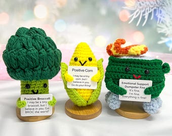 May include: Three crocheted vegetable figurines: a green broccoli, a yellow corn, and a green dumpster fire. Each holds a sign with positive messages. The set includes wooden bases and the text "3PCS+3Base".