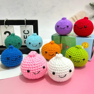 Könnte beinhalten: Eine Sammlung von neun bunten, gehäkelten Zwiebelspielzeugen mit lächelnden Gesichtern. Die Zwiebeln sind in verschiedenen Farben erhältlich, darunter Blau, Grün, Rosa, Orange, Weiß, Lila, Rot und Hellblau. Die Spielzeuge sind handgefertigt und wirken weich.