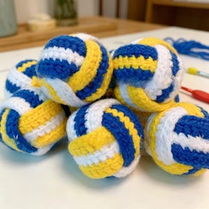 May include: A collection of small, spherical, crocheted items resembling volleyballs. Each sphere features a repeating pattern of thick blue, white, and yellow yarn stripes. The textured, handmade balls are clustered together on a flat surface.