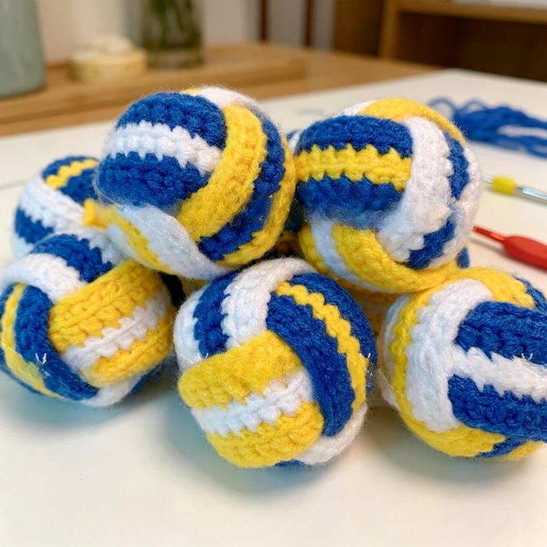 May include: A collection of small, spherical, crocheted items resembling volleyballs. Each sphere features a repeating pattern of thick blue, white, and yellow yarn stripes. The textured, handmade balls are clustered together on a flat surface.