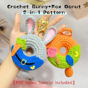 Könnte beinhalten: Zwei gehäkelte Donut-Schlüsselanhänger, ein graues Kaninchen und ein orangefarbener Fuchs, mit goldfarbenen Schlüsselringen. Der Kaninchen-Donut hat blaues Zuckerguss und eine Karotte, während der Fuchs-Donut grünes Zuckerguss und eine rosa Blume hat. Der Text lautet "Crochet Bunny+Fox Donut 2-in-1 Pattern".