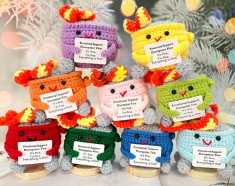 May include: Eight crocheted plush toys shaped like fire pits with smiling faces and holding signs that say "Emotional Support Dumpster Fire It's fine, I'm fine. Everything is fine!" The toys are in various colors, including purple, yellow, orange, pink, green, red, blue, and light blue.