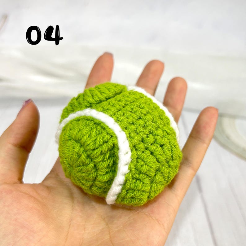 May include: A small, round, handmade crochet tennis ball featuring a vibrant lime green yarn texture and a distinct white yarn stripe pattern. The number 04 is printed in black in the upper left corner of the image, indicating the item label.