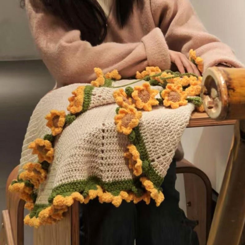K&ouml;nnte beinhalten: Eine geh&auml;kelte Decke mit Sonnenblumenmuster. Die Decke ist beige mit gr&uuml;nen und gelben Akzenten.