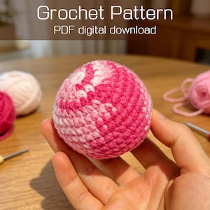 May include: A spherical crochet object featuring a variegated pattern of light pink, medium pink, and white yarn. The text "Crochet Pattern PDF digital download" is visible at the top. The object is held in a palm, with yarn balls and hooks nearby.