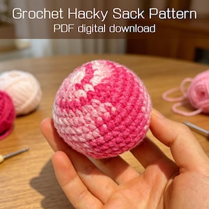 May include: A crocheted hacky sack in shades of pink and white, held in a hand. The text "Crochet Hacky Sack Pattern" and "PDF digital download" are visible at the top. Other balls of yarn and a crochet hook are in the background.