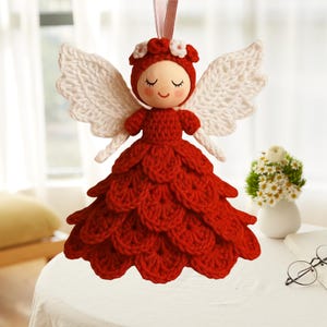 May include: A handmade crochet angel ornament with a red dress and white wings. The angel has a wooden face with closed eyes and a red bonnet with white flower accents. A pink ribbon is attached for hanging.