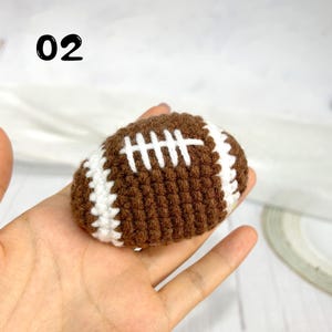 May include: A small, brown, crocheted American football with white stitched laces and white stripes at each end. The number 02 is printed in black in the upper left corner of the image, indicating a specific item in a collection.
