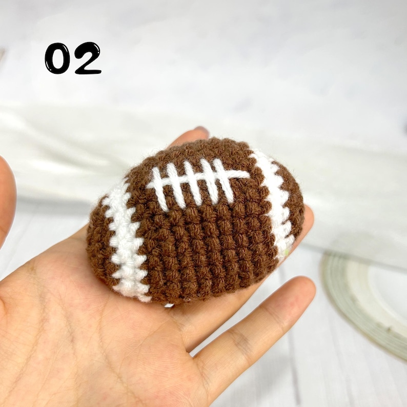 May include: A small, brown, crocheted American football with white stitched laces and white stripes at each end. The number 02 is printed in black in the upper left corner of the image, indicating a specific item in a collection.