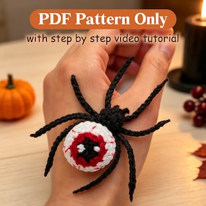 May include: A crochet spider with a white and red eyeball-shaped body and eight black legs. The text at the top reads PDF Pattern Only with step by step video tutorial. The spider features a central black pupil and red iris details.