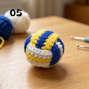 May include: A small, spherical, crocheted ball featuring a pattern of alternating stripes in bright yellow, royal blue, and white yarn. The textured, handmade item is round and compact, displaying clear, uniform stitches throughout its surface.