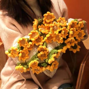 K&ouml;nnte beinhalten: Ein gestrickter Pullover mit Sonnenblumenmuster. Der Pullover ist wei&szlig; mit gr&uuml;nen Streifen und hat gelbe geh&auml;kelte Sonnenblumen.