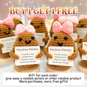 May include: A collection of crocheted potato figures, each with a smiling face, holding a sign that reads "Positive Potato." The potatoes have pink bows and are displayed on wooden bases. The text "BUY 1 GET 1 FREE" is at the top.