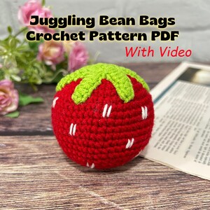 May include: A red crocheted strawberry-shaped bean bag with a green top and white stitched details. The text Juggling Bean Bags Crochet Pattern PDF With Video is displayed above the object. The item is round and textured with yarn stitches.
