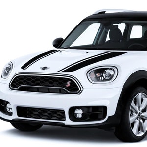 Peut inclure: Une Mini Cooper Countryman blanche avec des bandes de course noires sur le capot et le côté de la voiture. La voiture a un toit noir et des roues noires.