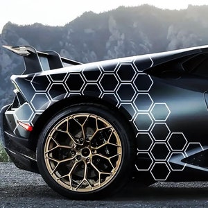 May include: A black Lamborghini with a white honeycomb pattern on the side. The car has gold wheels and is parked on a grey asphalt surface.