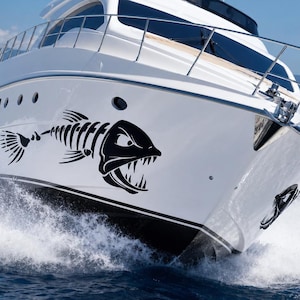 Könnte beinhalten: Eine weiße Motoryacht auf blauem Wasser mit einem großen schwarzen Grafikaufkleber eines stilisierten Fischskeletts mit scharfen Zähnen an der Rumpfseite. Das Schiff hat ein schlankes Design mit einer Metallreling und dunkel getönten Fenstern.