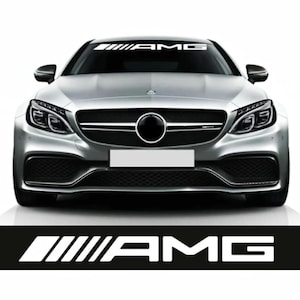 Op de afbeelding: Een zilveren auto met de witte tekst "AMG" bovenaan de voorruit. Een zwart-witte "AMG" sticker staat onderaan de afbeelding. De auto heeft een zwarte grille en koplampen.