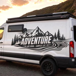 May include: A white camper van featuring a large black vinyl decal on the side. The graphic displays a mountain range, pine trees, and a winding river, with the text ADVENTURE OFF ROAD CAMPER LIFE printed in bold, capitalized white lettering.