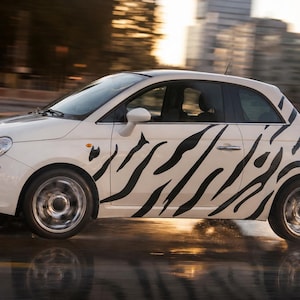 May include: A white two-door compact car featuring a black zebra stripe pattern along the side panels. The vehicle has rounded headlights, silver wheels, and a small antenna on the roof, captured in motion on a wet surface with blurred surroundings.