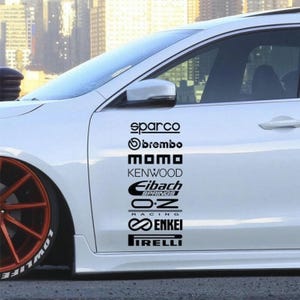 Puede incluir: Una puerta de coche blanca con una columna vertical de pegatinas negras de marcas automotrices que incluyen Sparco, Brembo, Momo, Kenwood, Eibach Springs, O.Z Racing, Enkei y Pirelli. El coche tiene una rueda naranja de radios múltiples.