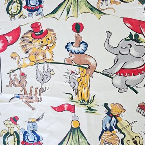 May include: A white fabric with a colorful circus theme print. The print features a lion, elephant, monkeys, a seal balancing a ball, a turtle on roller skates, a bird, and a tiger playing a cello. There are also two circus tents with green and yellow stripes.