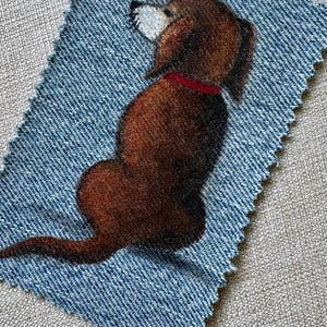 May include: A rectangular piece of blue denim fabric with pinked edges featuring a detailed illustration of a brown dachshund dog sitting and facing away. The dog has a white muzzle and wears a solid red collar around its neck, rendered in a soft texture.