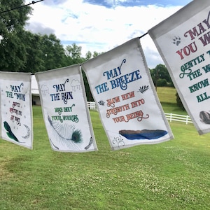 Peut inclure: Cinq drapeaux en tissu blanc avec du texte et des illustrations colorés. Les drapeaux sont suspendus à une ficelle et flottent au vent. Le texte sur les drapeaux se lit comme suit : "Que la lune vous restaure la nuit", "Que la pluie lave vos soucis", "Que la brise souffle une nouvelle force dans votre être", "Que vous puissiez marcher doucement à travers le monde et connaître sa beauté, tous les jours de votre vie".