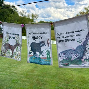 Op de afbeelding: Een reeks stoffen banners met illustraties over de natuur en tekst. De banners tonen afbeeldingen van een konijn, een hert, een wasbeer en opossums, met zinnen als "Mogen alle wezens vreedzaam zijn" en "Mogen alle wezens gelukkig zijn".