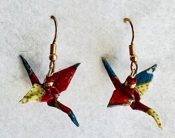 May include: A pair of origami crane earrings with gold hooks. The cranes are made of red, blue, and yellow patterned paper.