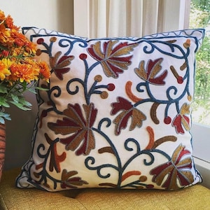 Op de afbeelding: Een wit kussen met een geborduurd bloemenmotief in tinten bruin, rood en blauw. Het kussen heeft ingewikkelde patronen van bladeren en wijnstokken.