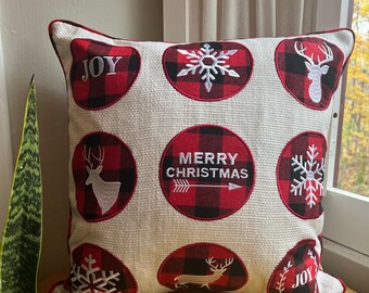 May include: Square decorative pillow with a cream-colored background and red and black plaid accents. The pillow features embroidered designs of snowflakes, deer, and the words "JOY" and "MERRY CHRISTMAS". The pillow has a red border.