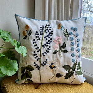 Embroidered floral pillow cover with a nature-inspired design. Features blue, pink, mustard, and green flowers and leaves on a cream linen background. The pillow is placed on a mustard seat by a window, with a green plant visible beside it.