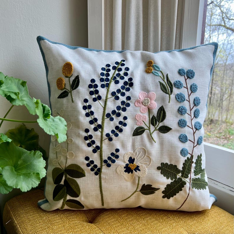 Embroidered floral pillow cover with a nature-inspired design. Features blue, pink, mustard, and green flowers and leaves on a cream linen background. The pillow is placed on a mustard seat by a window, with a green plant visible beside it.