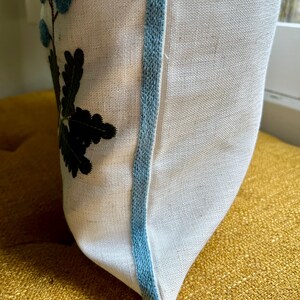 Puede incluir: Almohada blanca con ribete azul y hojas y flores negras bordadas.