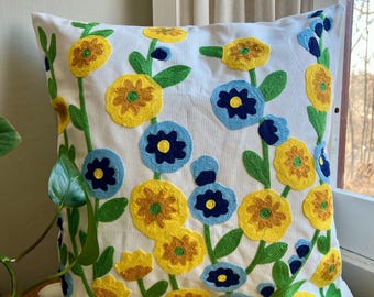 May include: White throw pillow with embroidered yellow, blue, and green flowers. The pillow is on a yellow surface.