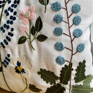 Puede incluir: Una almohada blanca con dise&ntilde;os florales bordados. La almohada presenta flores y hojas rosas, azules y verdes.
