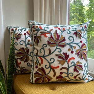 May include: Two white embroidered throw cushions with floral patterns in red, orange, gold, and teal. The square cushions rest on a yellow surface near a window. The embroidery features detailed leaf designs.