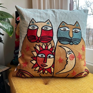 May include: Decorative square pillow with embroidered cat and celestial designs. The pillow features two stylized cats with red and blue faces, a sun, and a crescent moon. The background is a light blue and white checkered pattern.