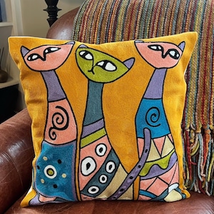 May include: Mustard yellow cushion featuring three stylized cat figures. The cats have pink, green, and blue details with geometric patterns and swirls. The cushion is on a brown leather chair.
