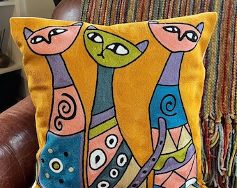 Peut inclure: Coussin décoratif jaune moutarde avec trois chats stylisés. Les chats ont des détails roses, verts et bleus avec des motifs géométriques et des volutes. Le coussin est sur une chaise en cuir marron.