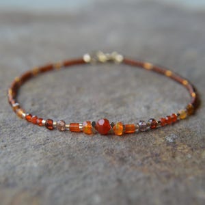 May include: A delicate bracelet featuring a mix of orange and amber-colored beads. The bracelet includes a central, round, orange gemstone and is secured with a gold-colored clasp. The beads vary in size and shape, creating a textured appearance.