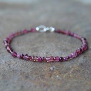 May include: A delicate bracelet featuring small, faceted pink gemstones strung together. The bracelet has a silver-colored clasp. The gemstones vary in shades of pink and magenta, creating a subtle gradient effect. The bracelet is designed to be worn around the wrist.