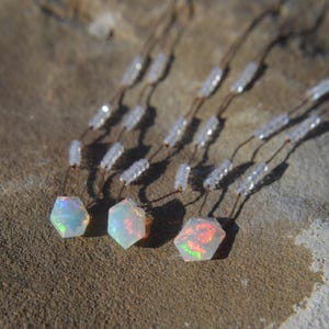 May include: Three hexagonal opal pendants with iridescent colors are displayed. Each pendant is suspended by a thin, brown cord, with a string of small, clear beads above the opal. The pendants are set against a textured, brown surface.