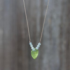 May include: A necklace featuring a carved green leaf-shaped pendant suspended from a thin, twisted beige cord. The pendant is accented by two small, pale blue faceted beads on each side, connecting the leaf to the cord in a V-shaped arrangement.