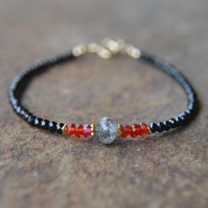 May include: A delicate bracelet featuring black beads, red gemstones, and a central gray stone. Gold-colored accents are interspersed throughout the design. The bracelet is displayed on a neutral-toned surface.